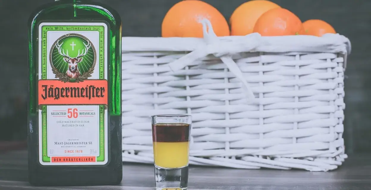 Butelka Jägermeistera i shot z dwoma warstwami, obok koszyk z pomarańczami. Idealne na wieczór, jak pić jagermeister.