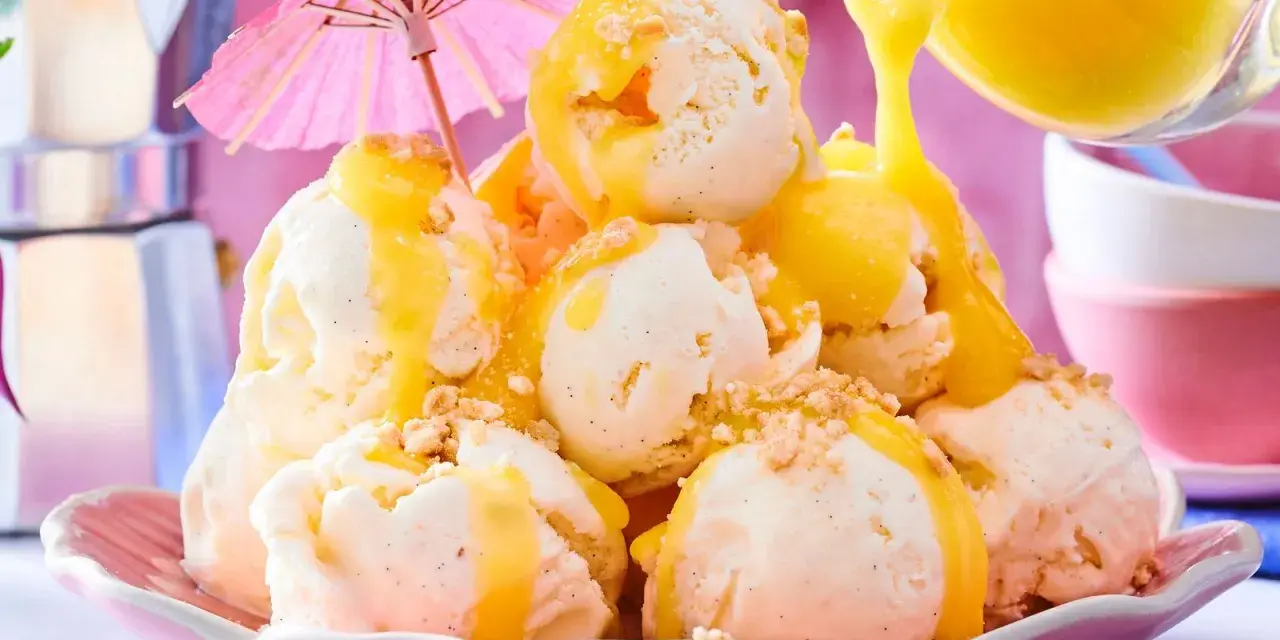 Ein Berg aus cremigem Vanilleeis, übergossen mit warmer Zitronensauce und bestreut mit Krümeln. Ein rosa Sonnenschirm sorgt für tropisches Flair.