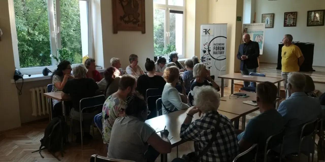 Spotkanie promujące język kaszubski. Ludzie siedzą przy stołach, słuchając prelegentów.