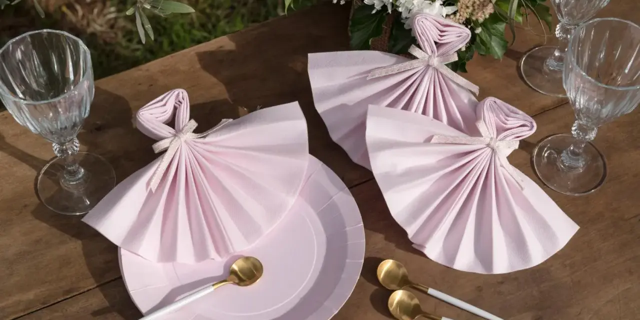 Vackert dukat bord med rosa servetter vikta som klänningar, guldfärgade skedar och glas. En bukett blommor fulländar dukningen.