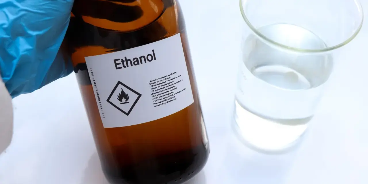 Braune Glasflasche mit der Aufschrift "Ethanol" und einem Warnsymbol. Daneben ein Becherglas mit klarer Flüssigkeit. Hier gibt es 100 prozentigen Alkohol.