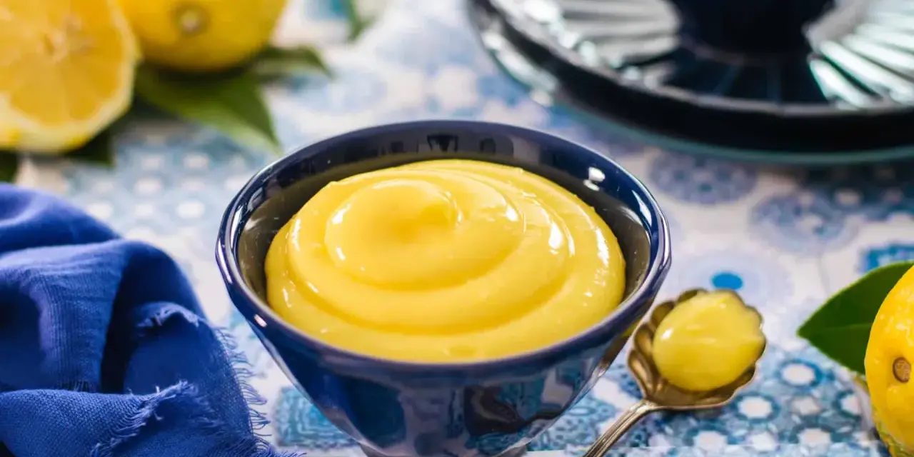 Una ciotola blu piena di vellutata crema al limone, con limoni freschi e un tovagliolo blu sullo sfondo.