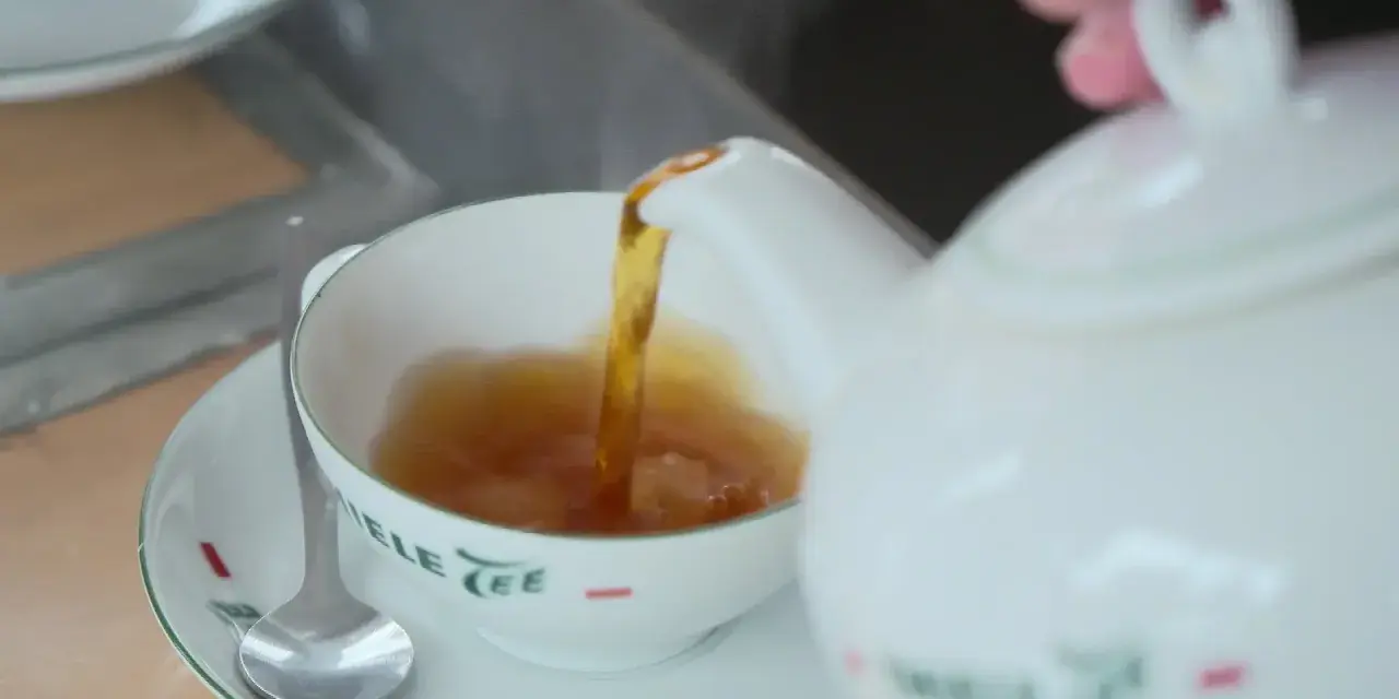 Tee mit Rum norddeutsch: Eine weiße Kanne gießt heißen Tee in eine Tasse. Ein Löffel liegt auf der Untertasse.