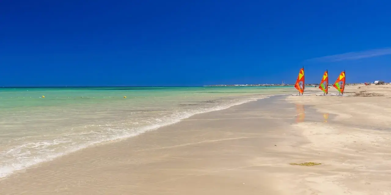 Plaża w Tunezji z trzema kolorowymi żaglami do windsurfingu na piasku i turkusową wodą.