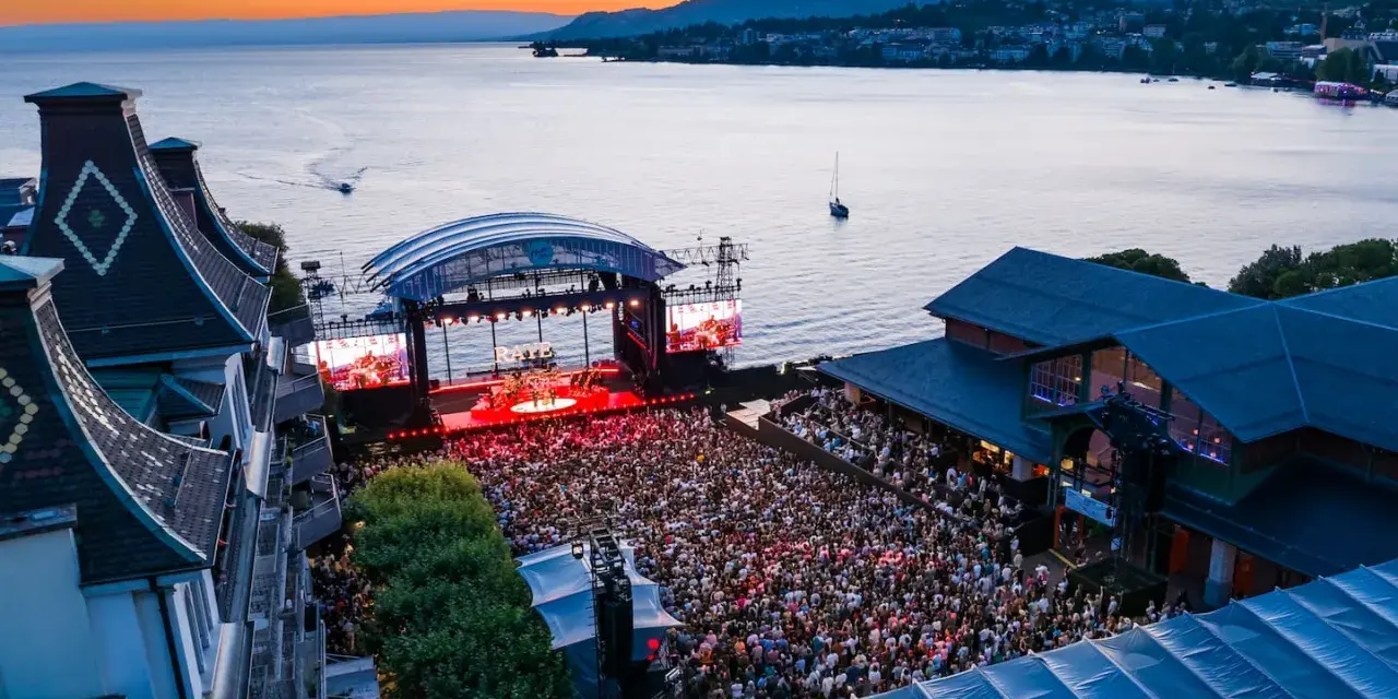 Ein großes Publikum feiert bei Sonnenuntergang ein Konzert am See. Die Bühne des Jazz Montreux Festivals ist beleuchtet, im Hintergrund der Genfersee.