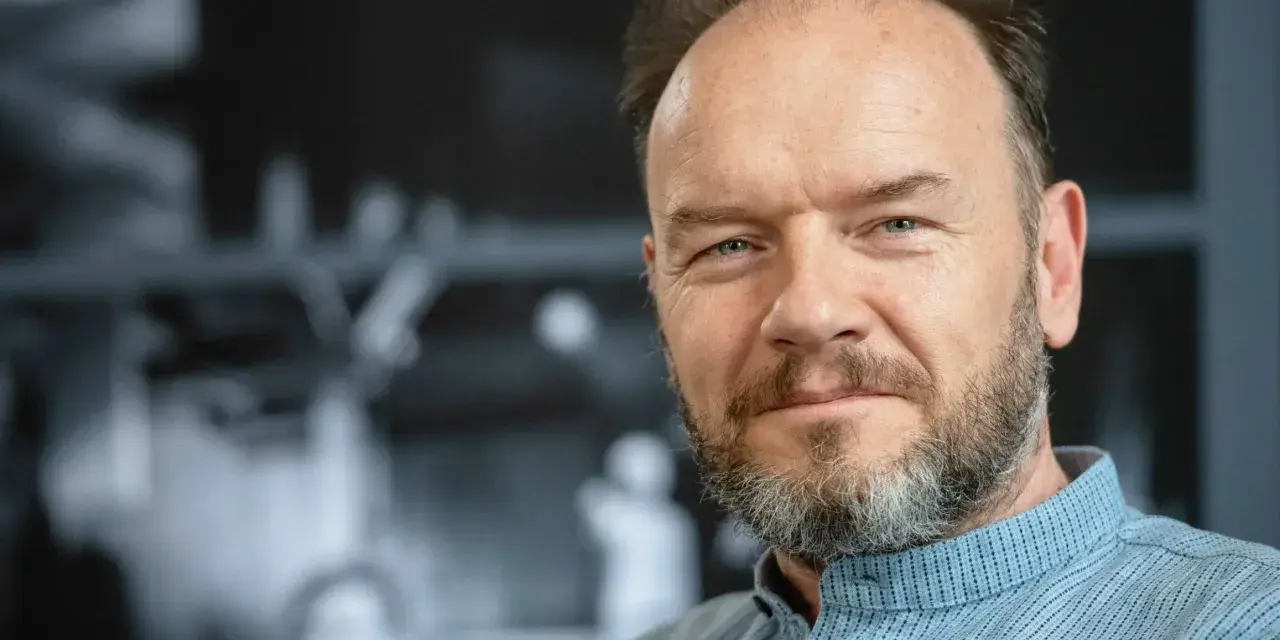 Portret mężczyzny z siwiejącą brodą, w niebieskiej koszuli. Redbad Klynstra dzieli się swoimi poglądami.
