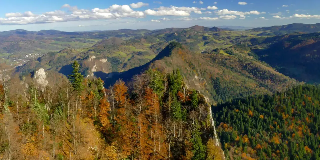 Jesień na szlaku Trzy Korony z Sromowiec Niżnych. Kolorowe drzewa na zboczu gór, w dole dolina i miasteczko.
