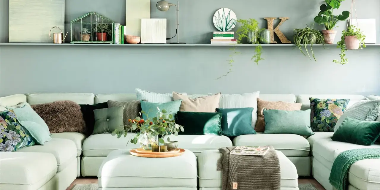 Sofá modular claro con cojines verdes y estampados, y una estantería con plantas y objetos decorativos, incluyendo una casita de cristal con **muebles pintados en verde**.