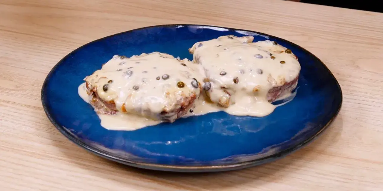 Due medaglioni di filetto al pepe verde, immersi in una cremosa salsa bianca, serviti su un piatto blu.