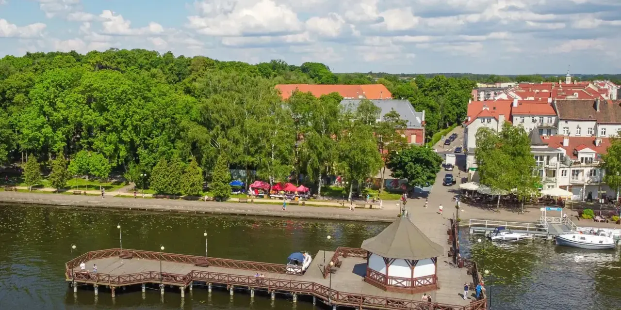 Idealne mazury na weekend: drewniane molo z altaną, jezioro, zielone drzewa i urokliwe miasteczko z czerwonymi dachami.