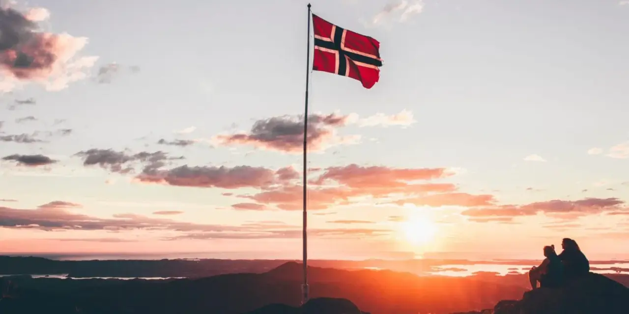 Zachód słońca w Norwegii. Dwie osoby siedzą na skale, patrząc na krajobraz. Flaga Norwegii powiewa na maszcie. Czy do Norwegii można lecieć na dowód osobisty?