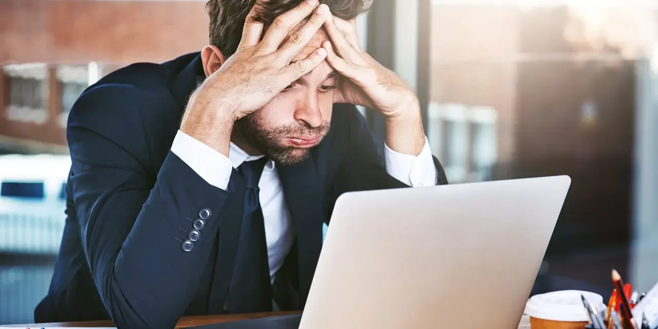 Zestresowany mężczyzna w garniturze z rękami na głowie, patrzący na laptopa. Trudna spłata długu po ogłoszeniu upadłości.
