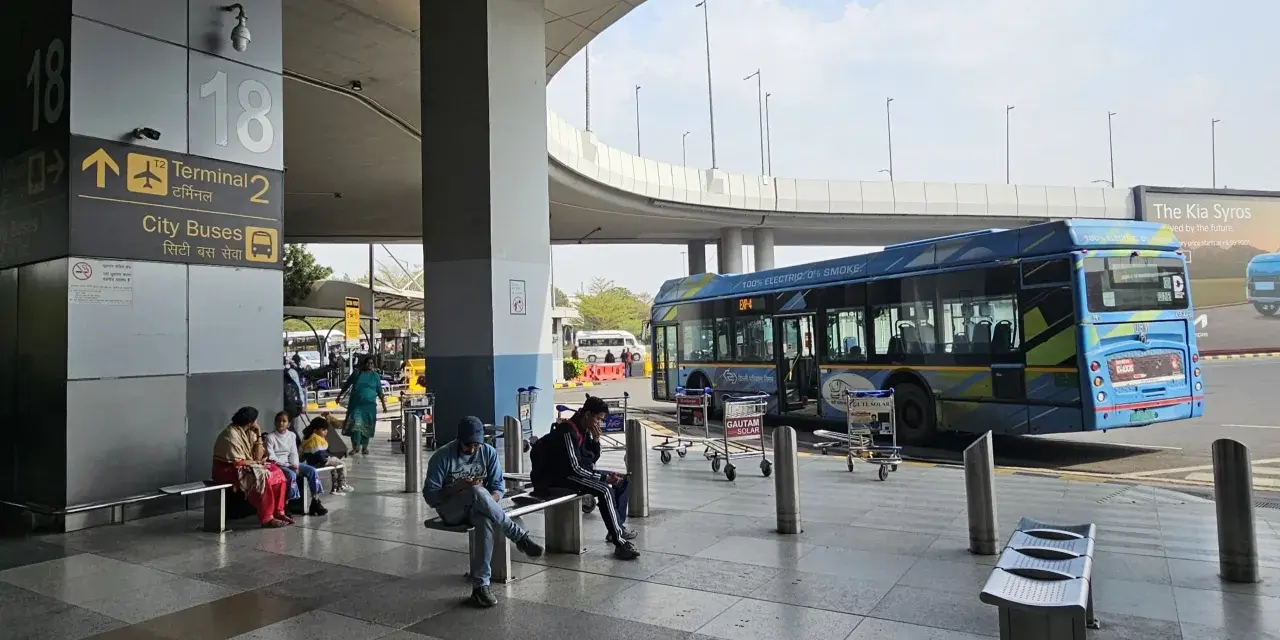 Oczekujący na autobusy na lotnisku Delhi. W tle niebieski autobus elektryczny.