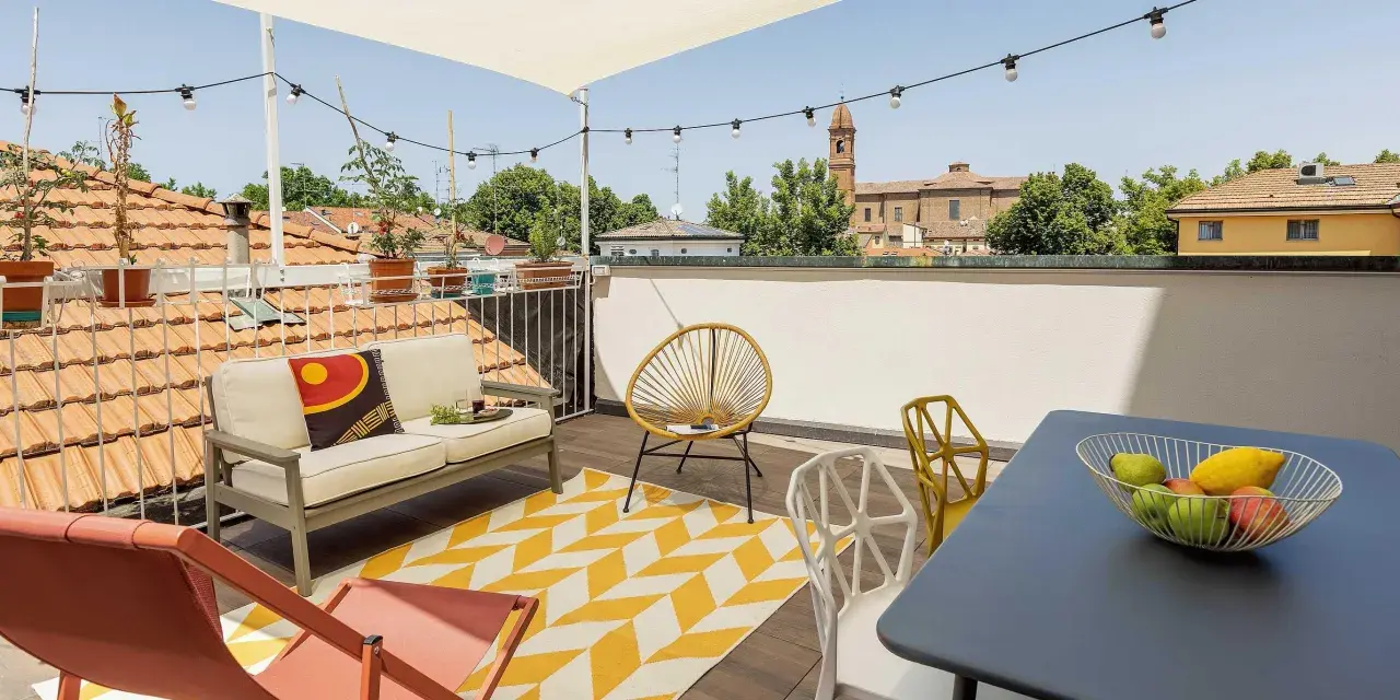Terrazza sul tetto idee: divano, sedie colorate, tappeto giallo e bianco, tavolino con frutta, luci e vista sui tetti.