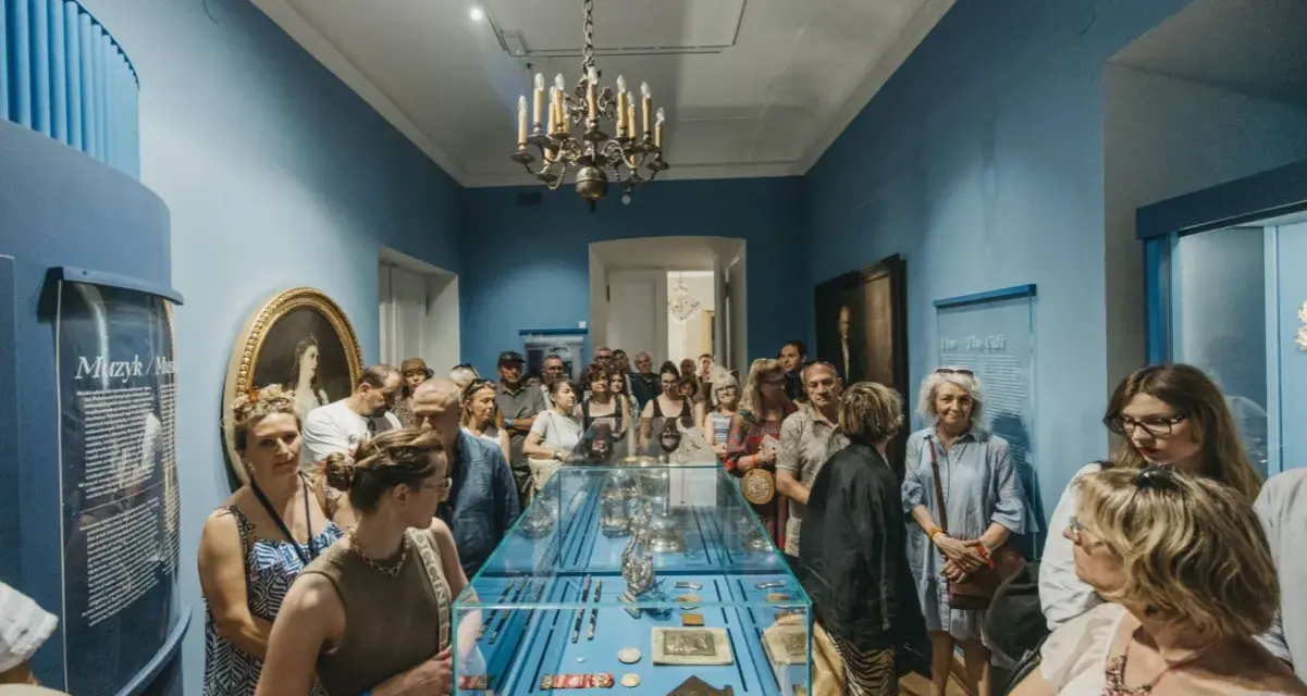 Grupa zwiedzających ogląda wystawę w muzeum. Wśród eksponatów w gablocie widać ozdobne przedmioty, a na ścianach obrazy i tablice informacyjne. Klasyczny wystrój sali z żyrandolem stanowi tło dla tego współczesnego zainteresowania historią.