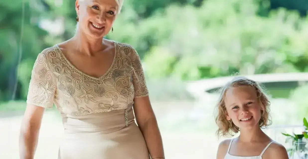 Elegancka stylizacja na komunię dla babci. Uśmiechnięta kobieta w beżowej sukni z haftem i dziewczynka w bieli.