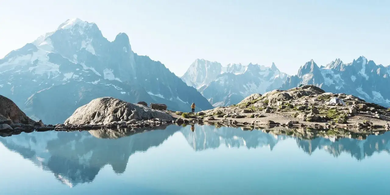 Un lac alpin reflète des montagnes majestueuses, un aperçu des plus belles randonnées de France. Une personne se tient sur la rive rocheuse.
