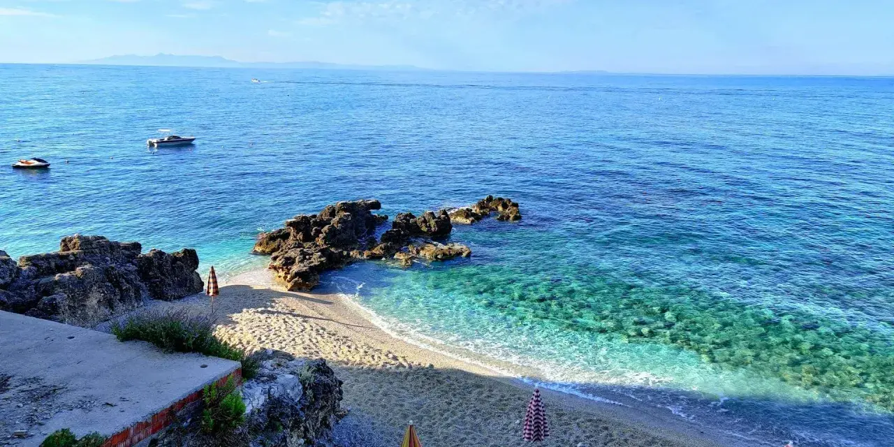Albania, czerwiec: słoneczna pogoda nad krystalicznie czystym morzem, kamienista plaża z parasolami i łódkami na horyzoncie.