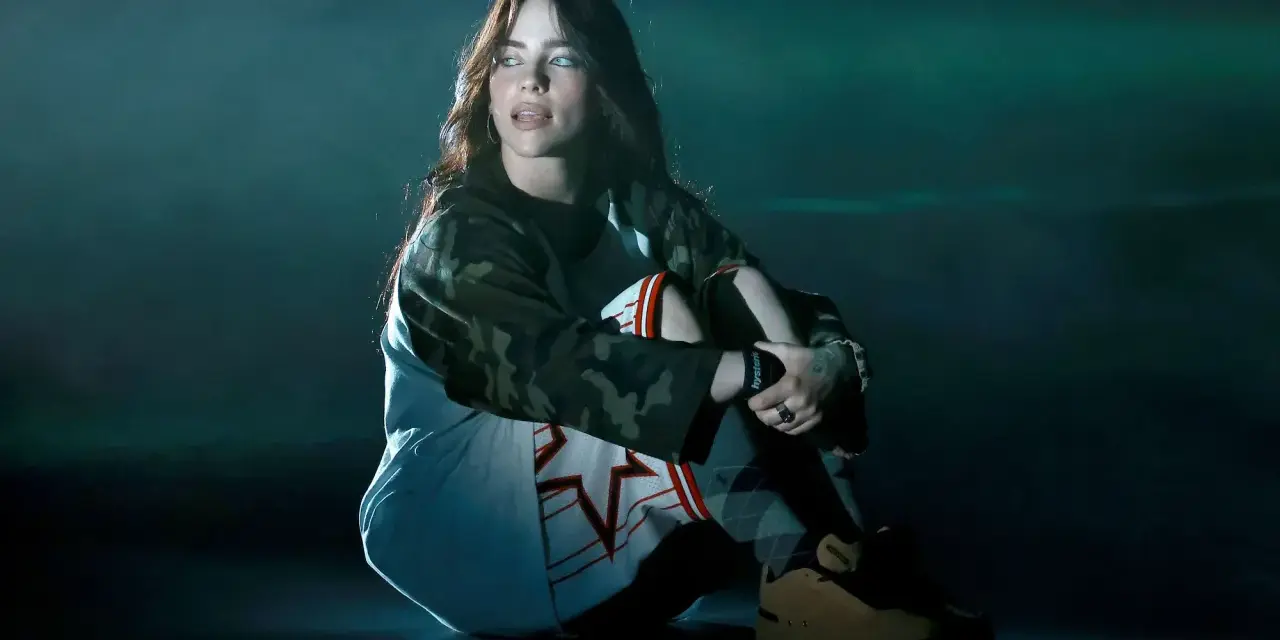 Billie Eilish siedzi na scenie, wpatrzona w dal. Jej stylizacja nawiązuje do estetyki jej albumu.