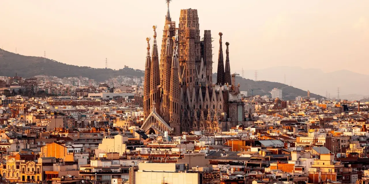 Vue panoramique de Barcelone au coucher du soleil, avec la Sagrada Familia au centre. Les travaux de la Sagrada Familia continuent, sa silhouette imposante se détachant sur le ciel orangé.