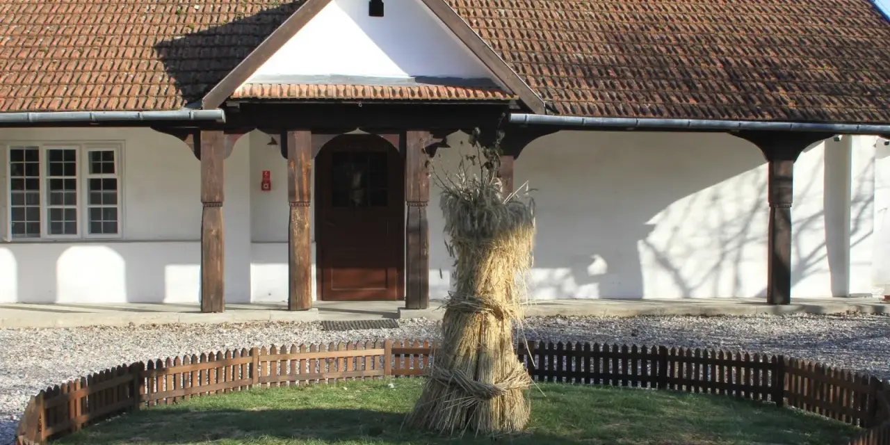 Bronowicka chata symbol: snop zboża przed tradycyjnym domem z drewnianymi elementami i ceramicznym dachem.