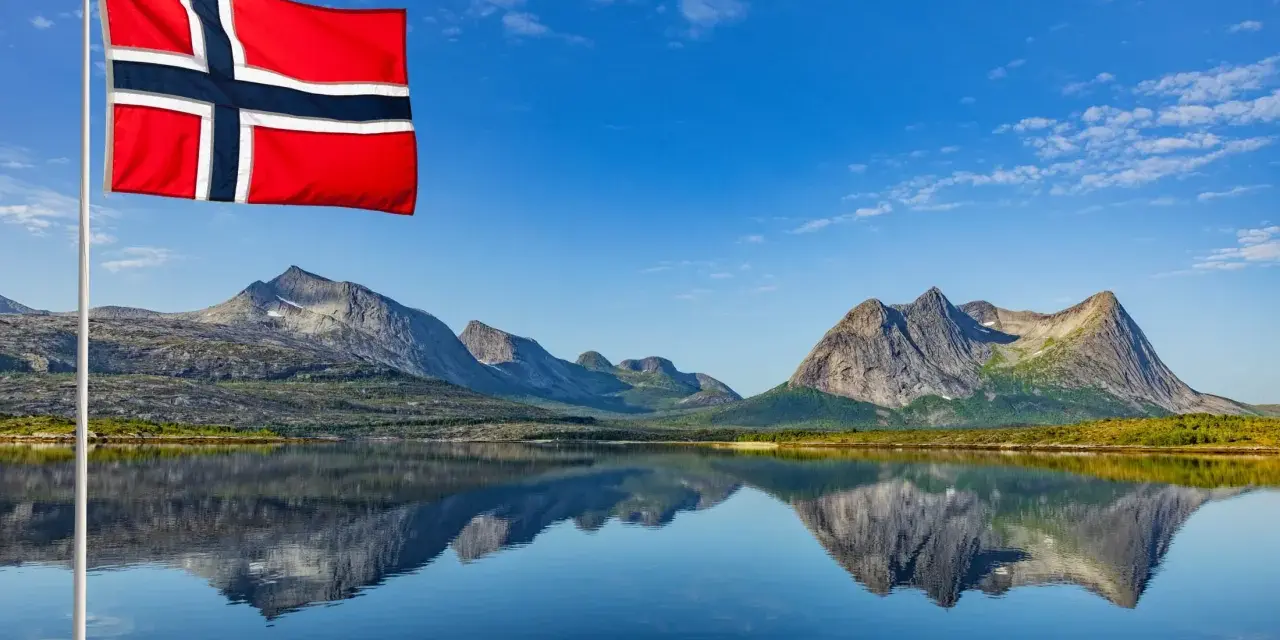 Norweska flaga powiewa nad jeziorem, odbijając się w wodzie. Czy do Norwegii potrzebny jest paszport? Piękne góry w tle.