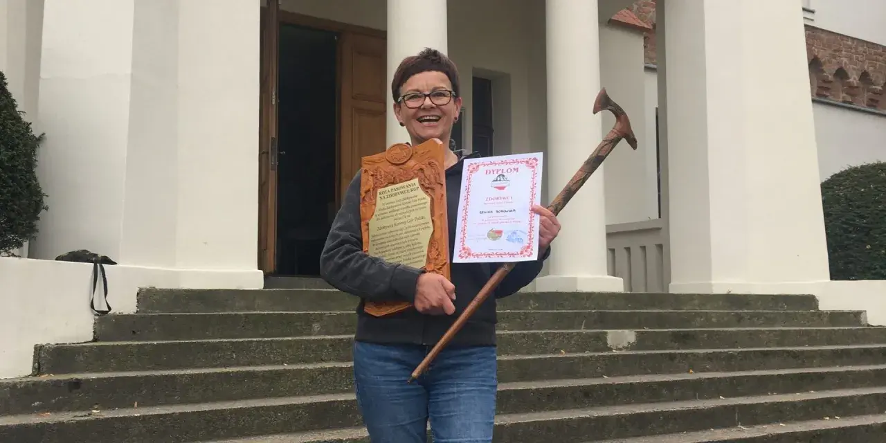 Uśmiechnięta kobieta z dyplomem i drewnianą laską, zdobywczyni Korony Gór Polskich.
