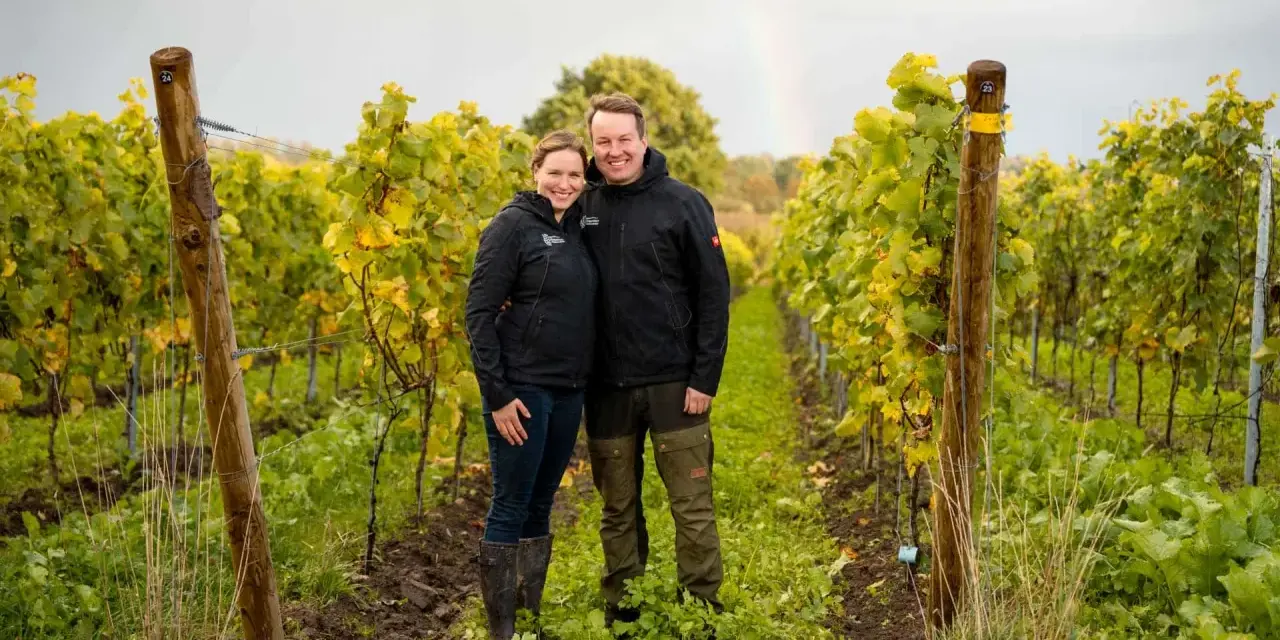 Ein Paar steht lächelnd in einem Weinberg in Schleswig-Holstein. Im Hintergrund ist ein Regenbogen zu sehen.
