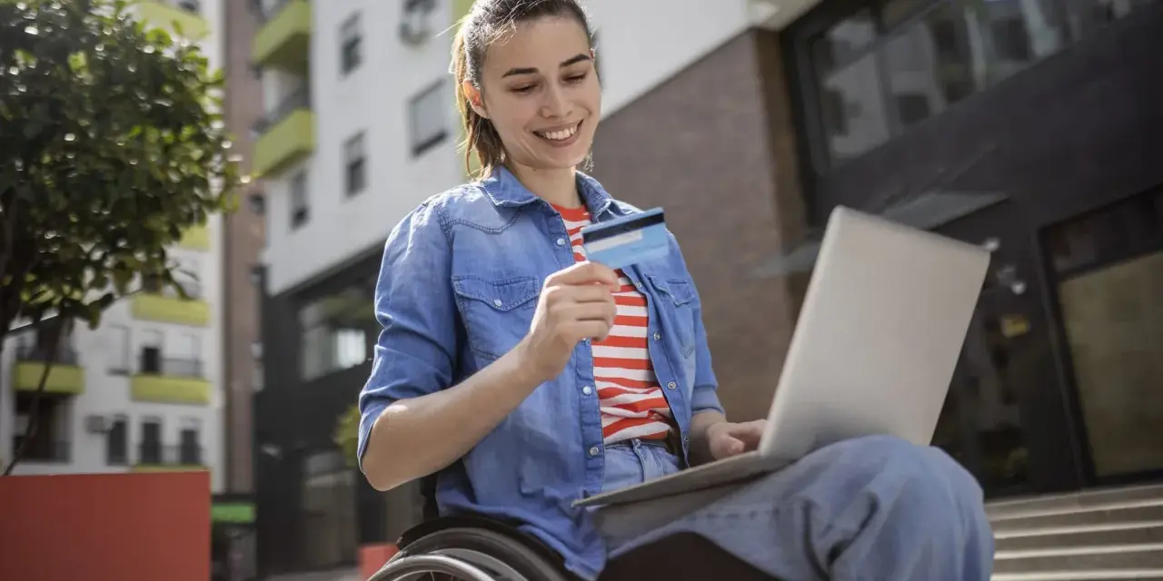 Uśmiechnięta kobieta na wózku inwalidzkim, korzystająca z laptopa i karty płatniczej, pokazuje, że stopnie niepełnosprawności nie ograniczają jej aktywności.