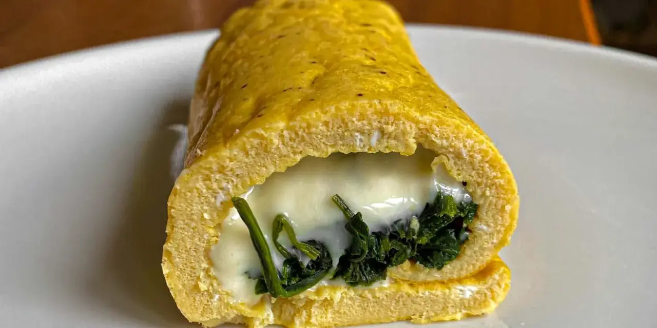 Rotolo di frittata dorato, ripieno di spinaci e formaggio filante, servito su un piatto bianco.