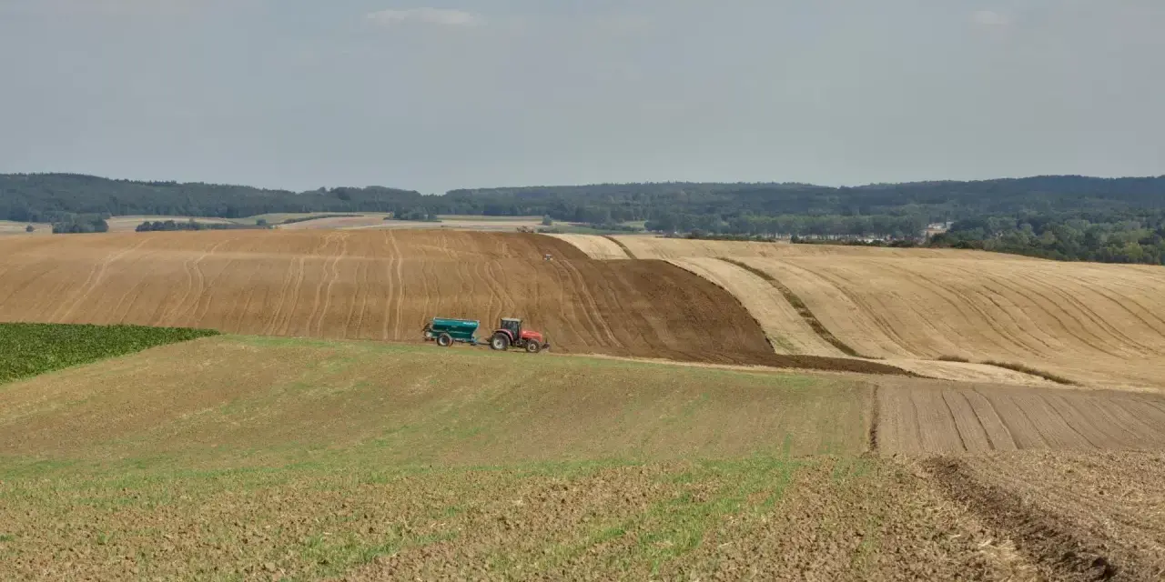 Czerwony traktor z przyczepą nawozi pole, przygotowując je do dalszej rekultywacji. W tle widać kolejne pola i las.