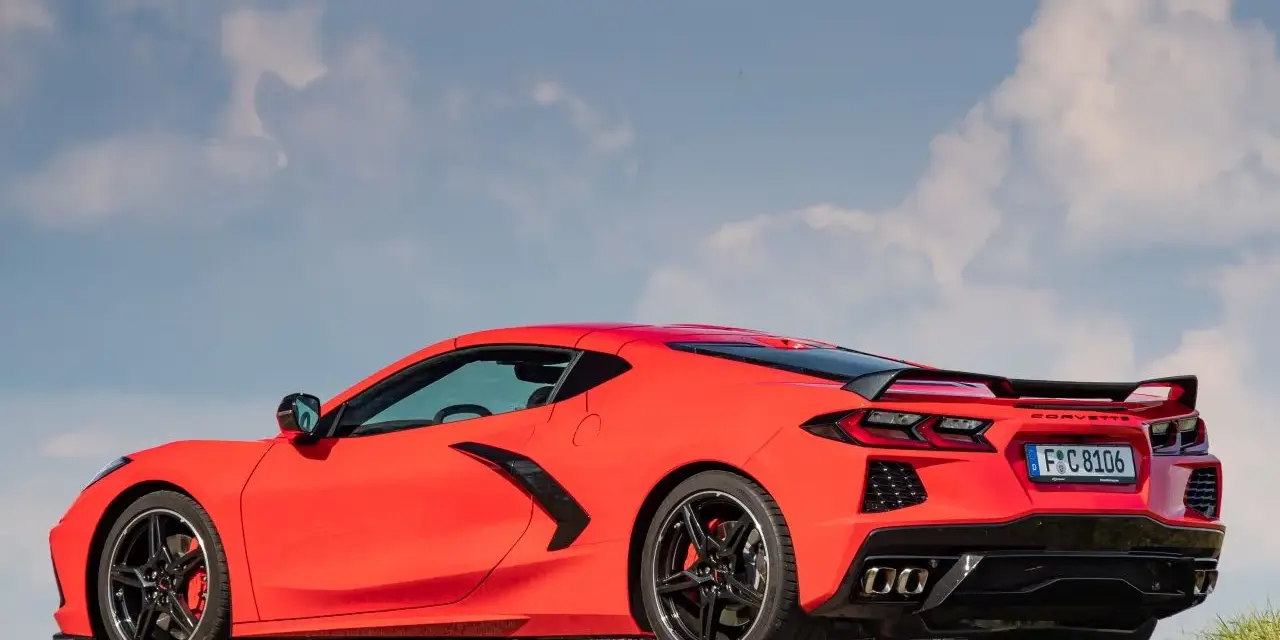 Czerwony Chevrolet Corvette C8 z czarnym spoilerem i felgami, zaparkowany na drodze pod błękitnym niebem z chmurami.