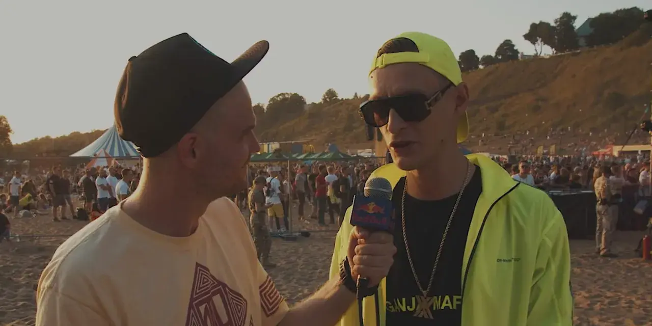 Dwóch mężczyzn na plaży, jeden z mikrofonem Red Bull, w tle tłum ludzi i scena.