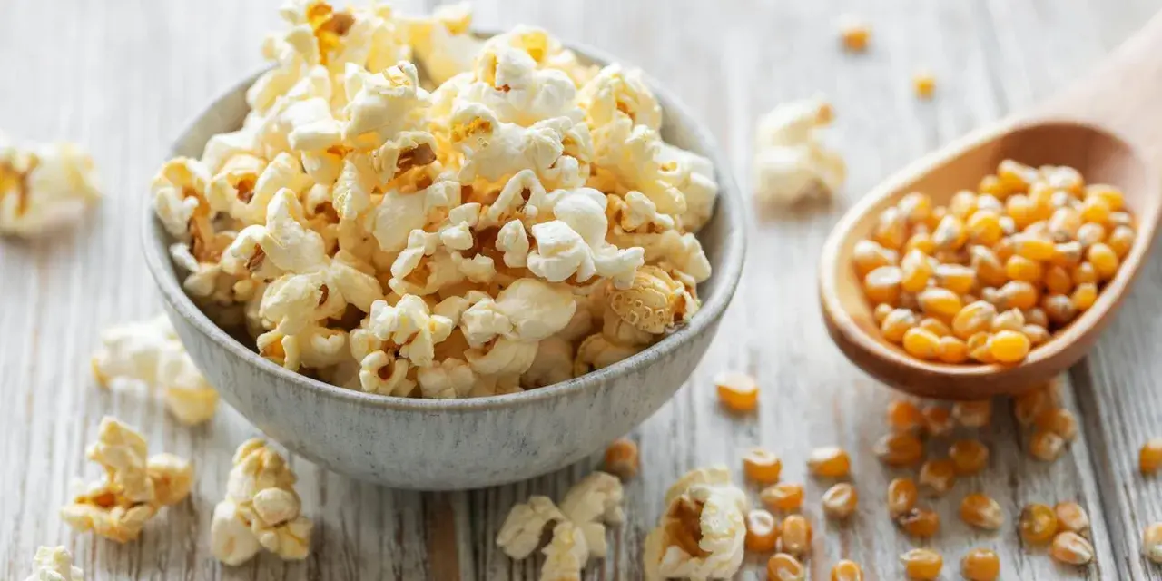 Schüssel mit frisch zubereitetem Popcorn im Topf, daneben eine Holzschaufel voller Maiskörner.