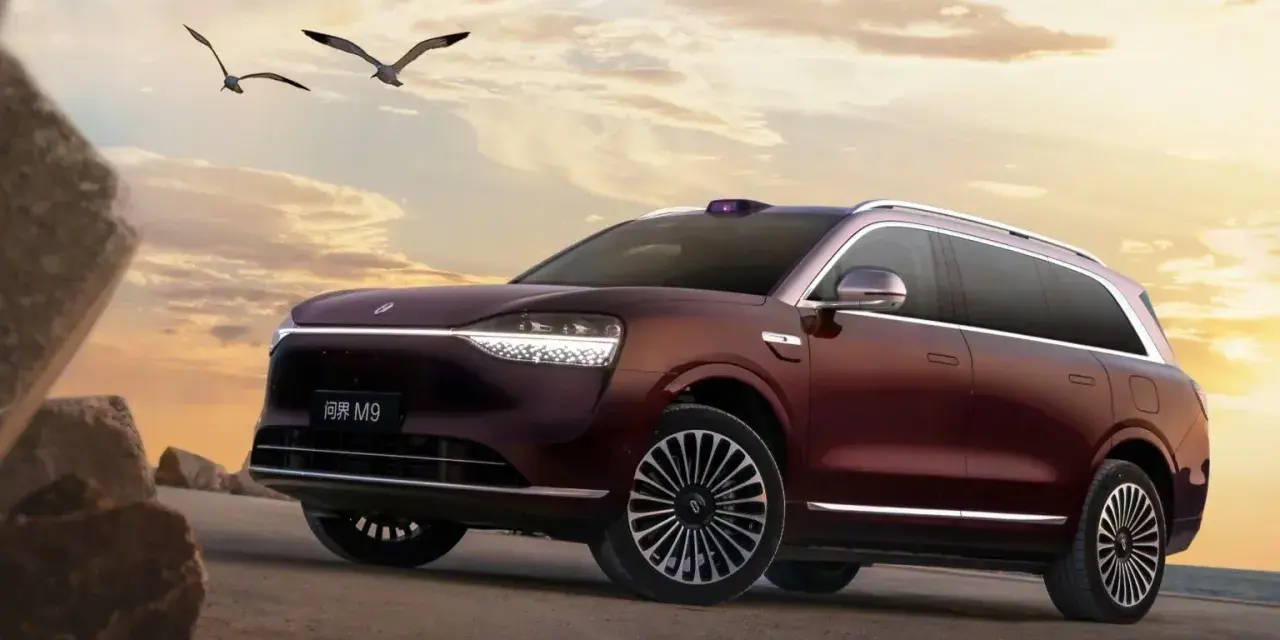 Ciemnoczerwony SUV Aito M9 na tle zachodzącego słońca, z ptakami w locie.