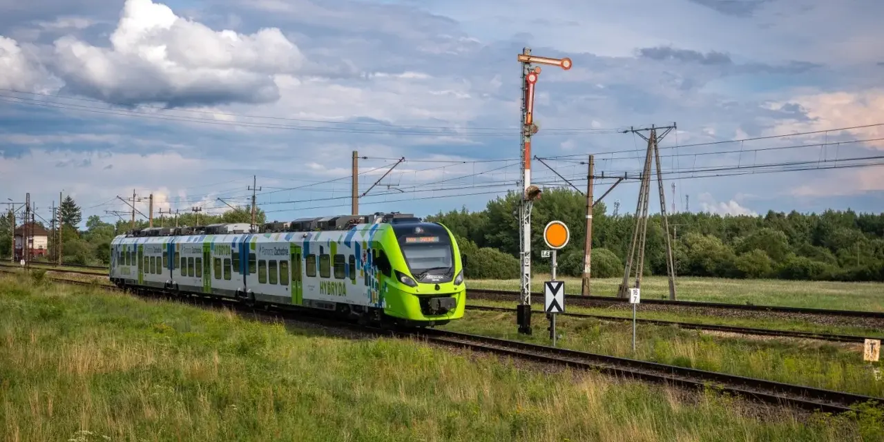 Zielony pociąg hybrydowy PKP jedzie po torach wśród zielonych pól. W tle las i chmury.
