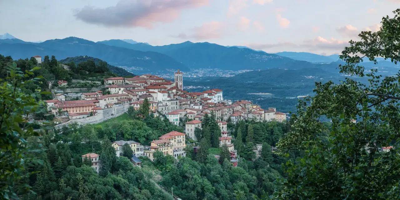 Borgo pittoresco su un colle, con case dai tetti rossi e un campanile, incastonato tra montagne e alberi. Via Francisca del Lucomagno, un incanto.