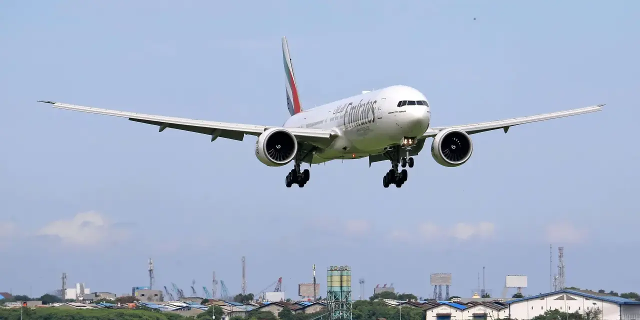 Emirates Boeing 777 zbliża się do lądowania. W tle widać przemysłowy krajobraz i inne samoloty na niebie, sugerujące ruch lotniczy, być może związane z dżerba loty.