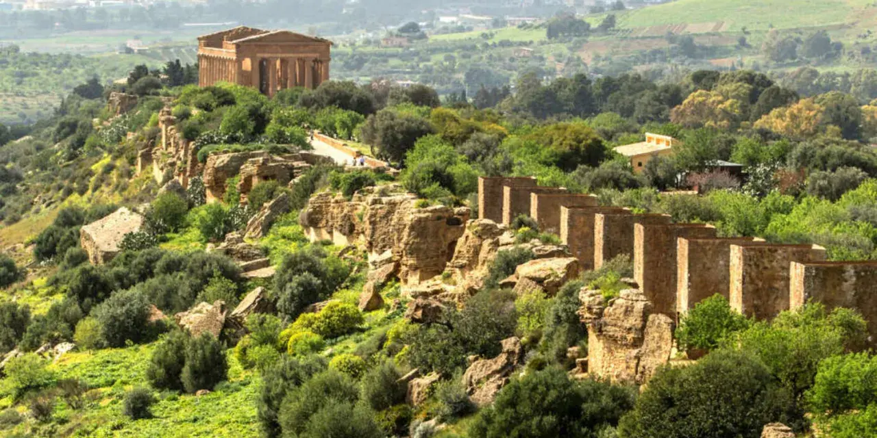 Ancient Greek temples stand proudly on a ridge in the Valley of the Temples, Sicily, surrounded by lush greenery and a distant town.