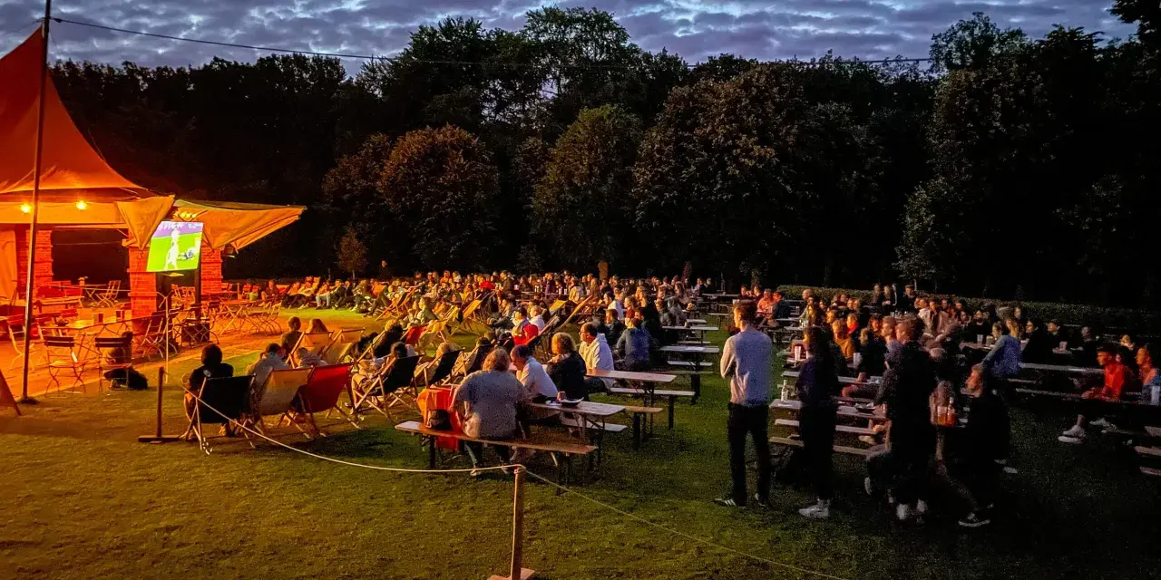 Menschen genießen ein Fußballspiel beim Public Viewing in Berlin unter einem dramatischen Abendhimmel.
