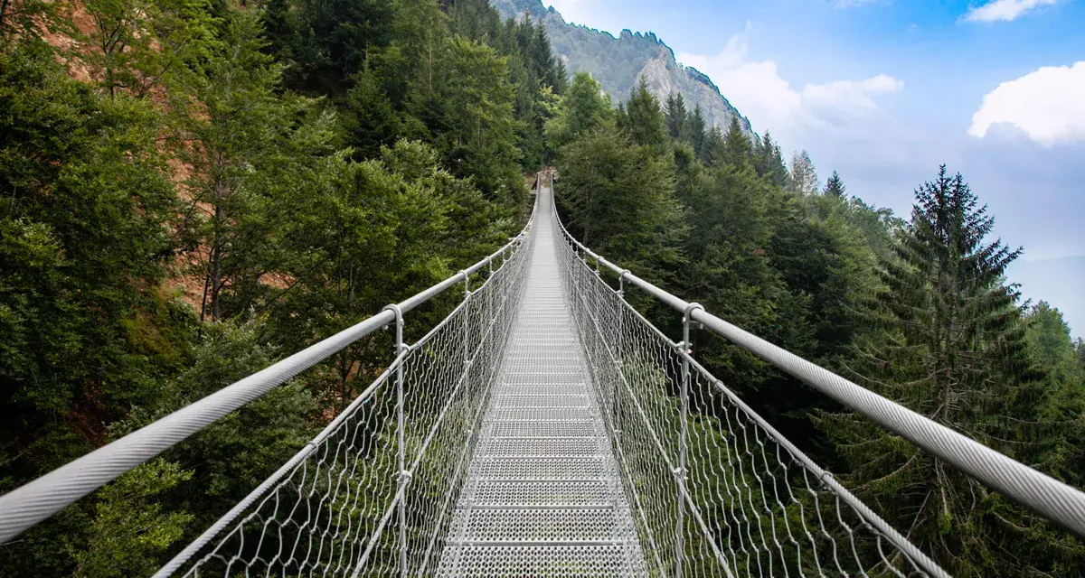 Un lungo ponte tibetano originale attraversa una foresta lussureggiante, con montagne sullo sfondo.
