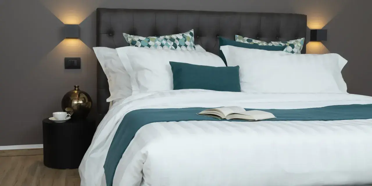 Letto rifatto con lenzuola bianche e copriletto turchese. Un libro aperto è posato al centro, pronto per essere letto. Ecco come si mette il lenzuolo di sopra.