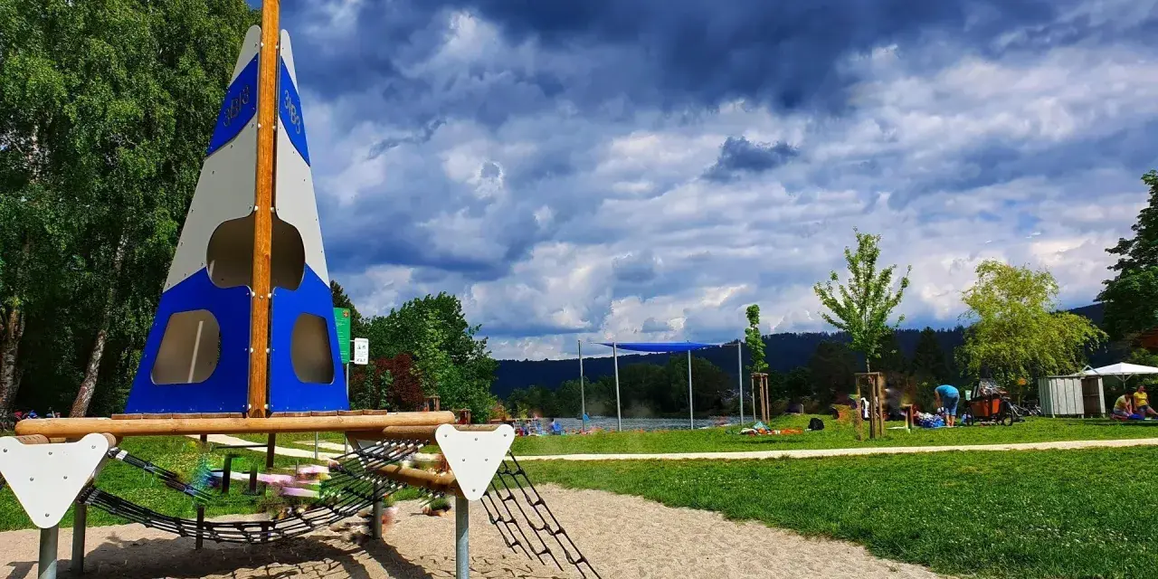 Spielplatz am Badesee Ludwigsburg mit Segel-Klettergerüst und Sandkasten. Im Hintergrund der See und Menschen.