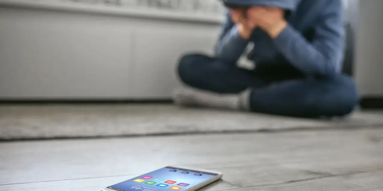 Osoba w kapturze siedzi na podłodze, ukrywając twarz w dłoniach. Obok leży telefon, symbolizujący izolację i dystymię.