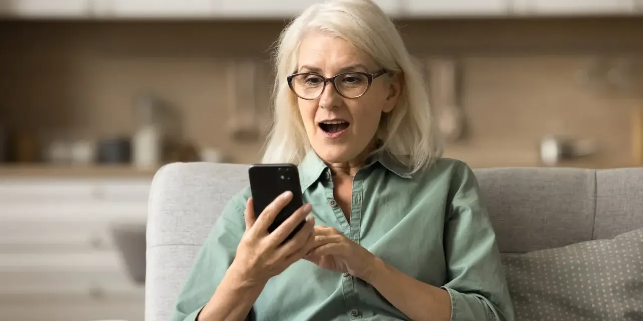 Zaskoczona kobieta z telefonem w dłoni dowiaduje się o zasiłki przedemerytalne po nowemu.