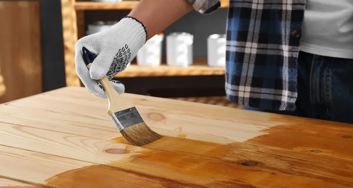 Main d'une personne portant un gant blanc appliquant du vernis sur une table en bois avec un pinceau. La peinture sur bois vernis prend forme.