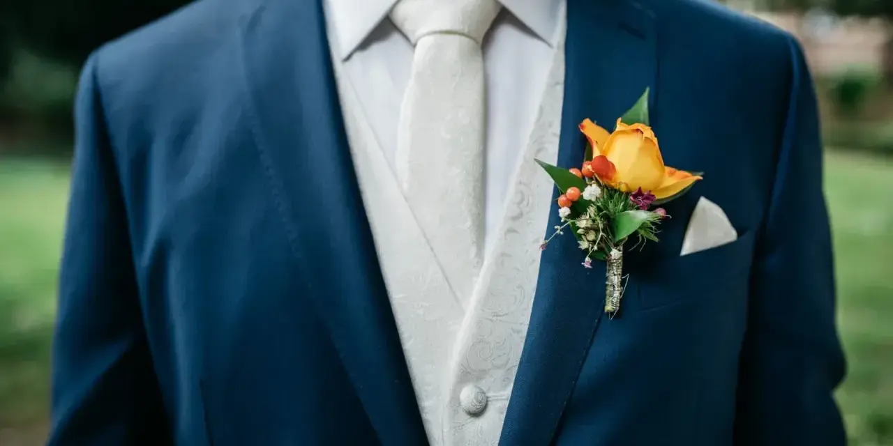 Elegancki, granatowy garnitur na wesele z białym, wzorzystym kamizelką i krawatem. Na klapie marynarki ozdobna butonierka z żółtą różą.