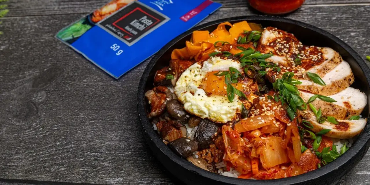 Pyszny Bibimbap przepis: ryż, kurczak, jajko sadzone, grzyby, marchewka i kimchi. Obok opakowanie pasty kimchi.