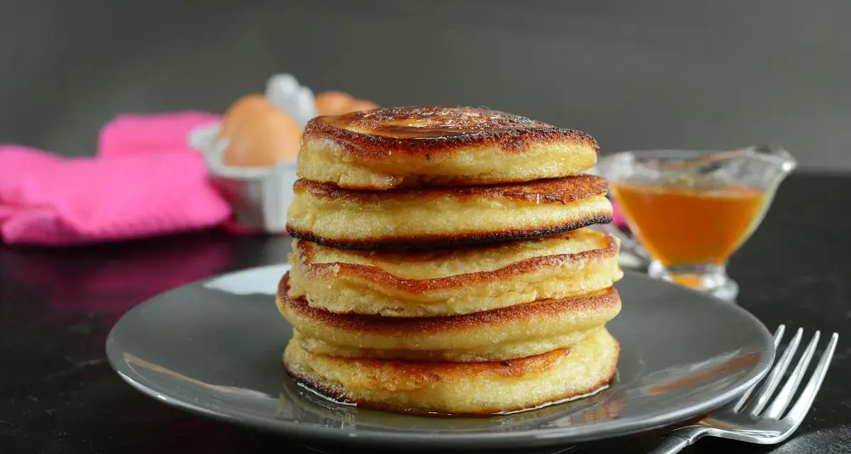 Pila di pancake senza latte, dorati e soffici, con sciroppo d'acero e uova sullo sfondo.