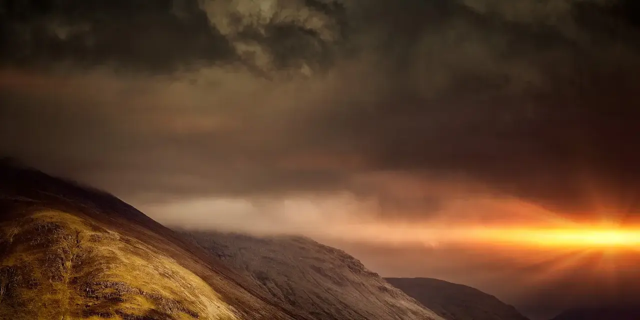 Dramatic sunburst pierces dark clouds over a rugged mountain range, a powerful display of the sovereignty of God.